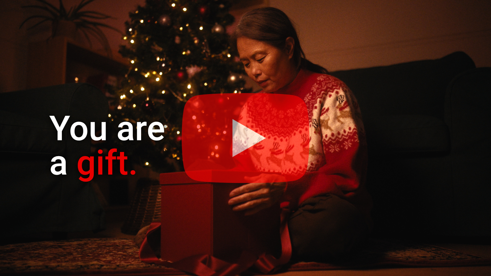 A women sits in front of a Christmas tree opening a present.