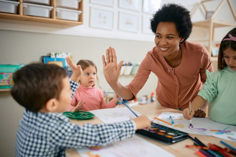 An adult giving a high five to a child, and other children painting around a table.