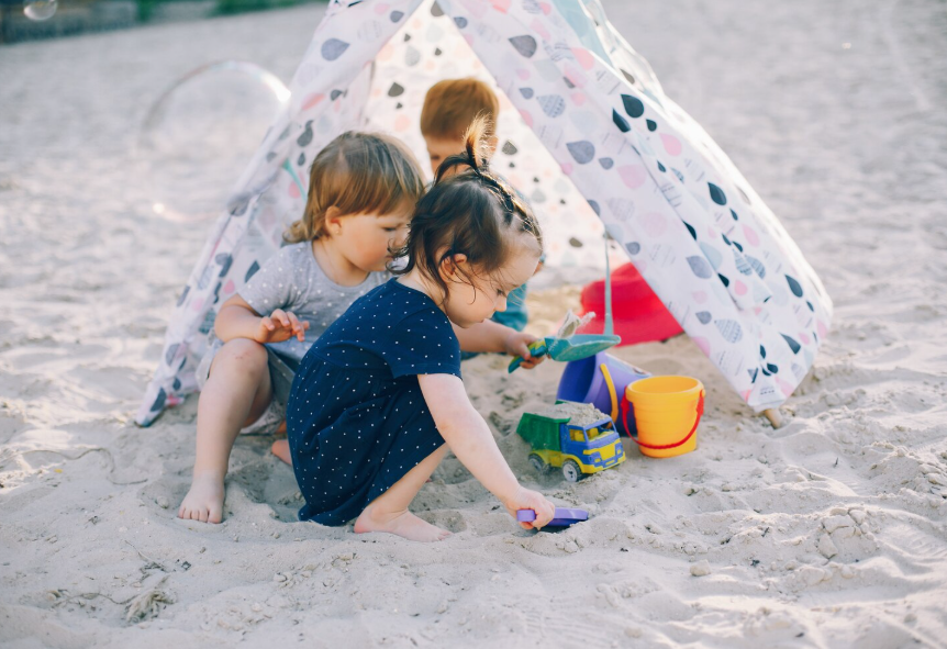 Three young children playing in sand near a small patterned play tent with buckets and spades