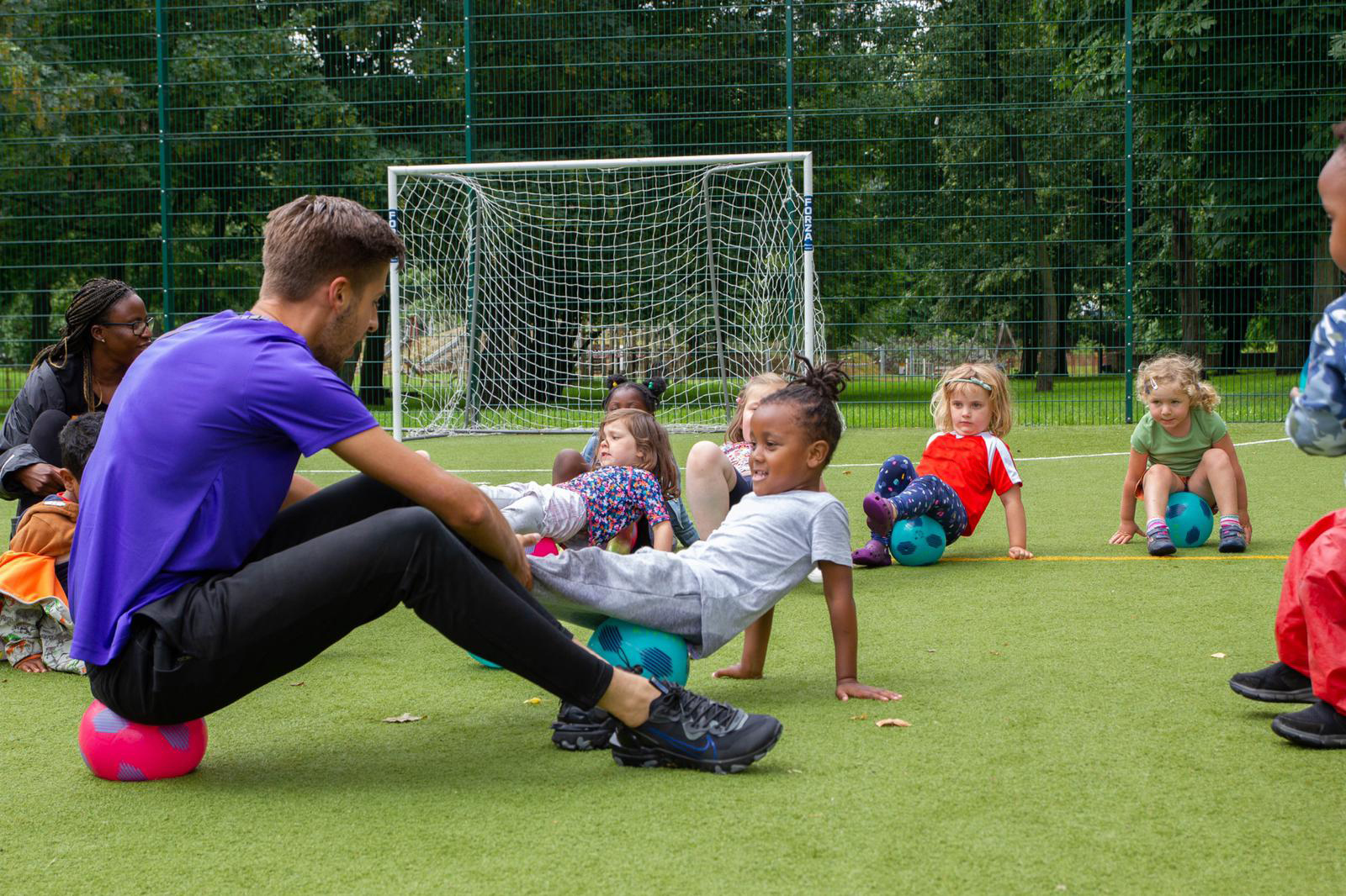 A person sitting on a ball with a group of young children each sitting on a ball with their hands placed on the floor behind their backs.