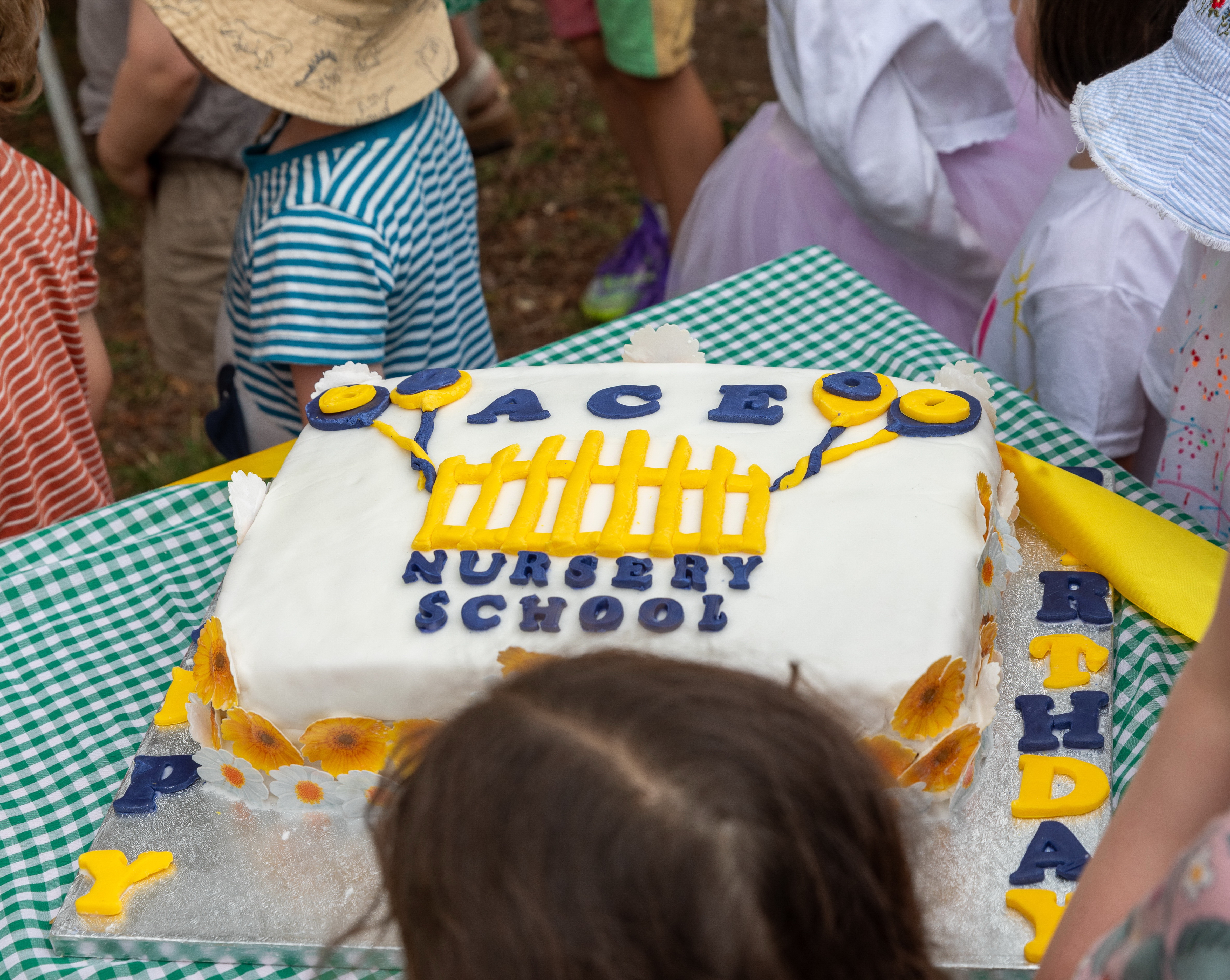 Photograph of a rectangular birthday cake decorated with white icing, yellow and blue balloons, and a yellow fence design, featuring the text "ACE NURSERY SCHOOL." Children wearing colourful clothing and hats surround the cake on a green and white checkered tablecloth, indicating a celebratory event at a nursery school.