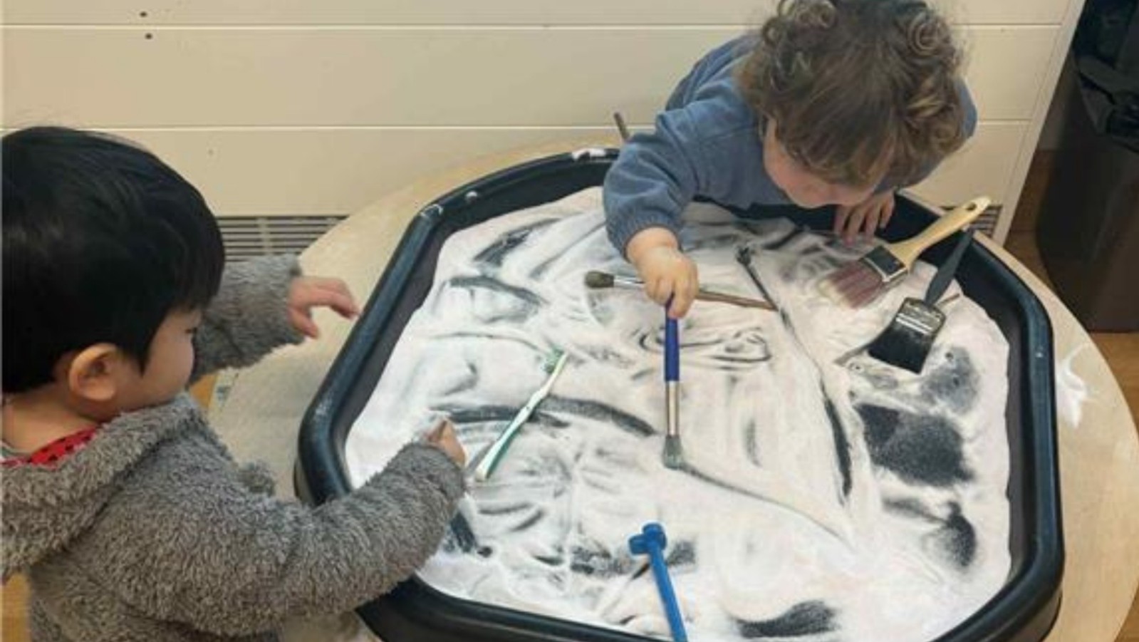 Two young children playing with brushes in a large black tray filled with white sand