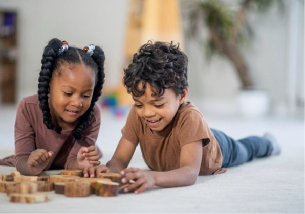 Two children lying on a floor, engaging with wooden blocks arranged in front of them.