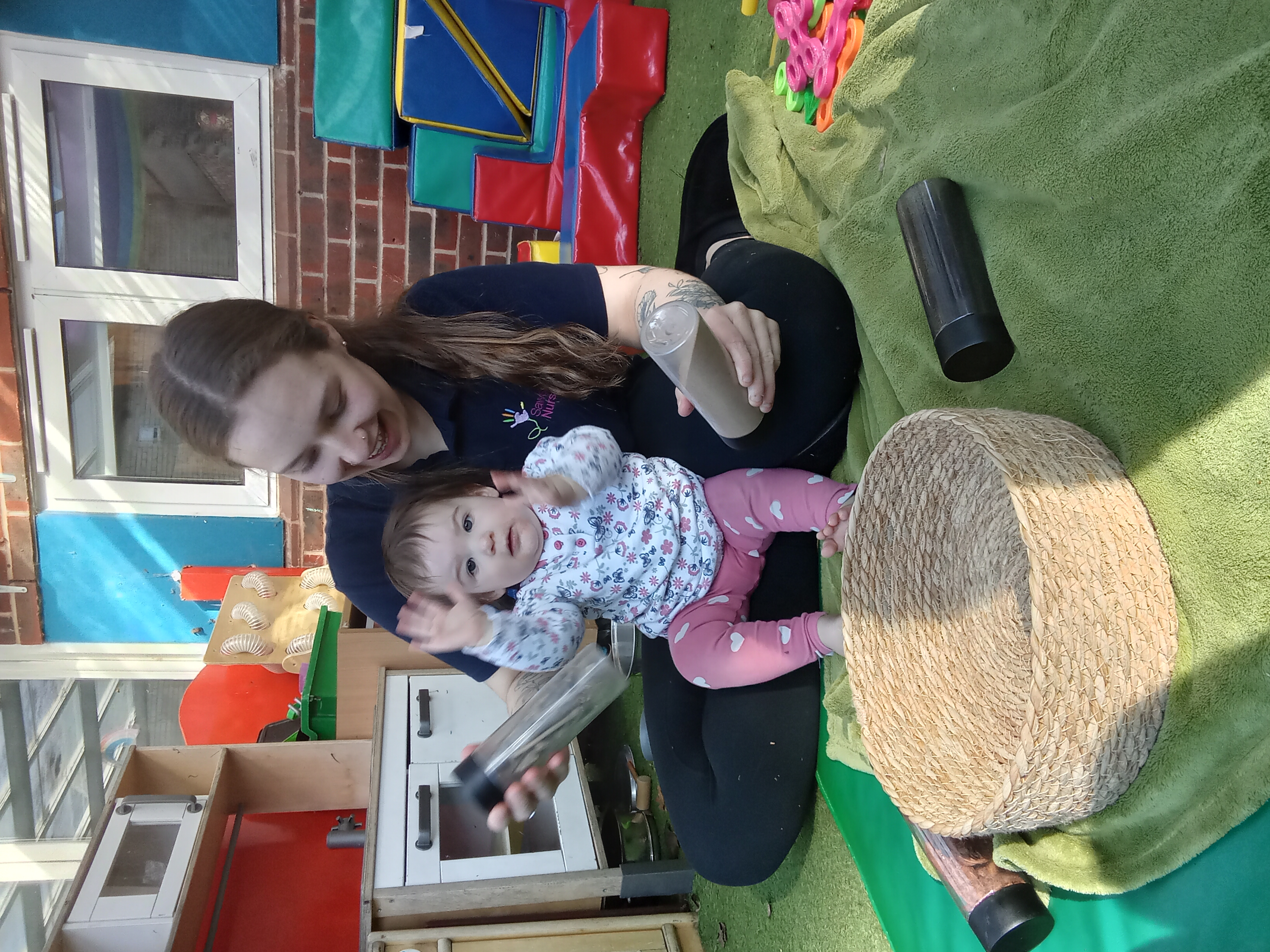 Photograph of a person and a baby sitting on a green blanket in a colourful playroom, engaging with musical instruments like a shaker and a rainstick. The setting includes a wicker basket, foam blocks, and bright toys, highlighting a playful and interactive moment.