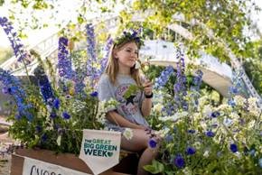 A person sitting among tall purple and white flowers in a garden setting with a white arched structure in the background. A sign reading "Great Big Green Week" is visible, indicating participation in an environmental or gardening event.