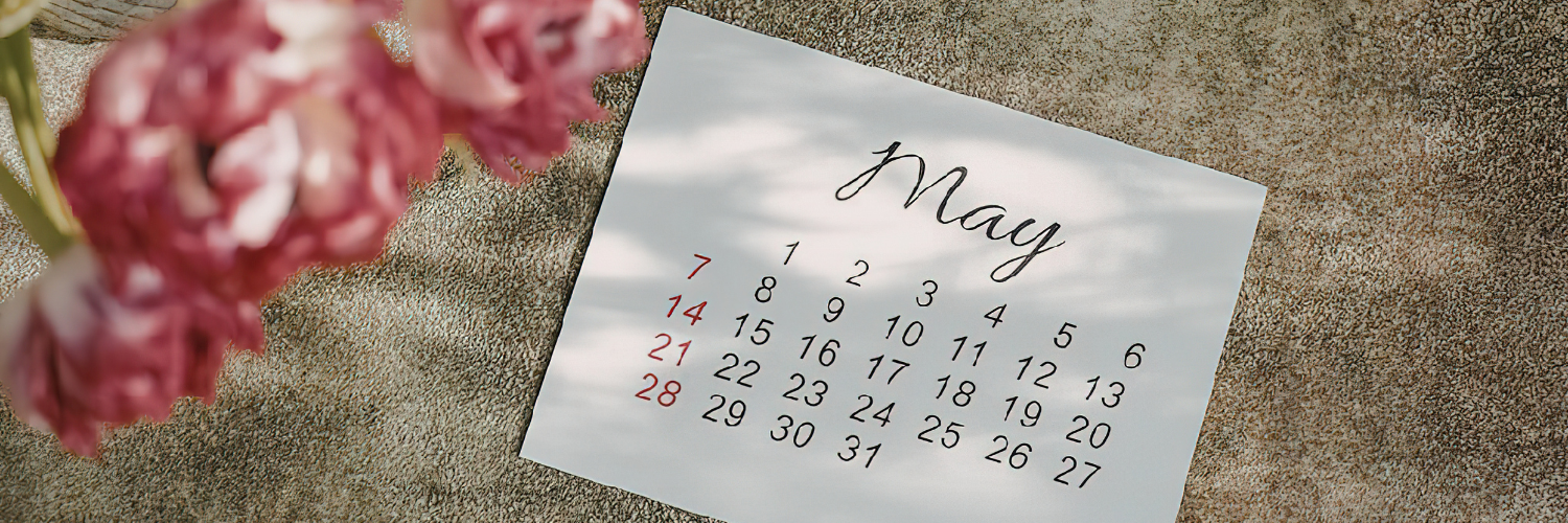Image shows a calendar open on the month of May laying on a table next to a bunch of pink flowers.