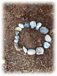 A photograph shows a circle made of variously sized and coloured stones arranged on dry, brown soil. A metal rod or stick lies near the top of the circle, suggesting an outdoor setting or a nature-based activity