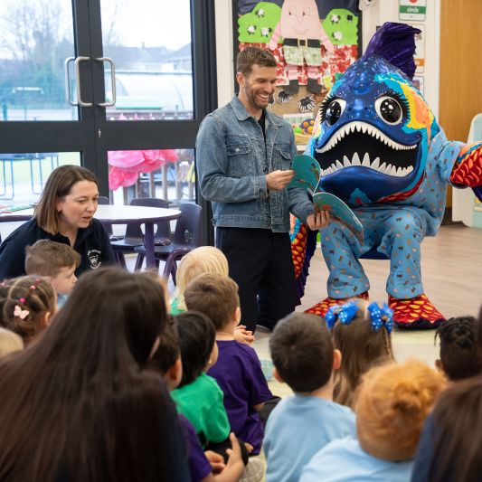 A group of young children seated on the floor, attentively watching a person in a colourful, large fish costume with big eyes and sharp teeth, who appears to be engaging them in an educational or entertainment activity. The setting is a bright indoor classroom or library with windows, child-sized furniture, and colourful decorations on the walls.
