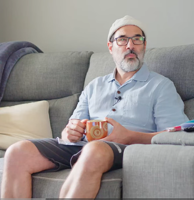 Adam Buxton sitting on a gray couch holding a patterned mug, wearing a light blue button-up shirt and dark shorts