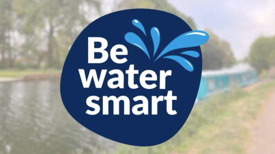 A photograph featuring a dark blue water droplet-shaped graphic with white text "Be water smart" and blue water splash illustrations. The background shows a blurred canal scene with trees and a narrowboat, emphasizing water conservation awareness.