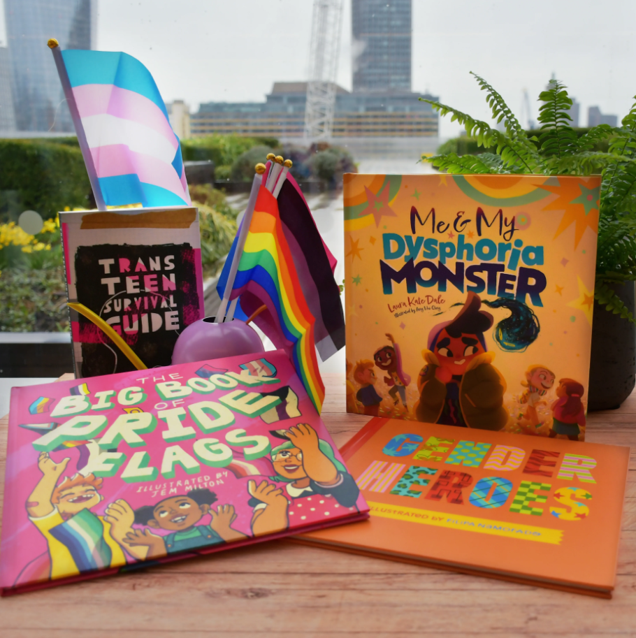 Photograph of a colourful display featuring LGBTQ+ themed books and flags arranged on a table with a cityscape background. Books include "Trans Teen Survival Guide," "Me & My Dysphoria Monster," "The Big Book of Pride Flags," and "Gender Heroes," accompanied by transgender and rainbow pride flags, highlighting themes of identity, support, and celebration.