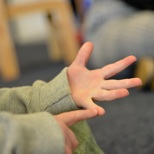Photograph shows close-up hands in grey sweaters, one palm extended with fingers spread while another finger points toward it. Blurred indoor background emphasizes nonverbal communication or gesture demonstration.