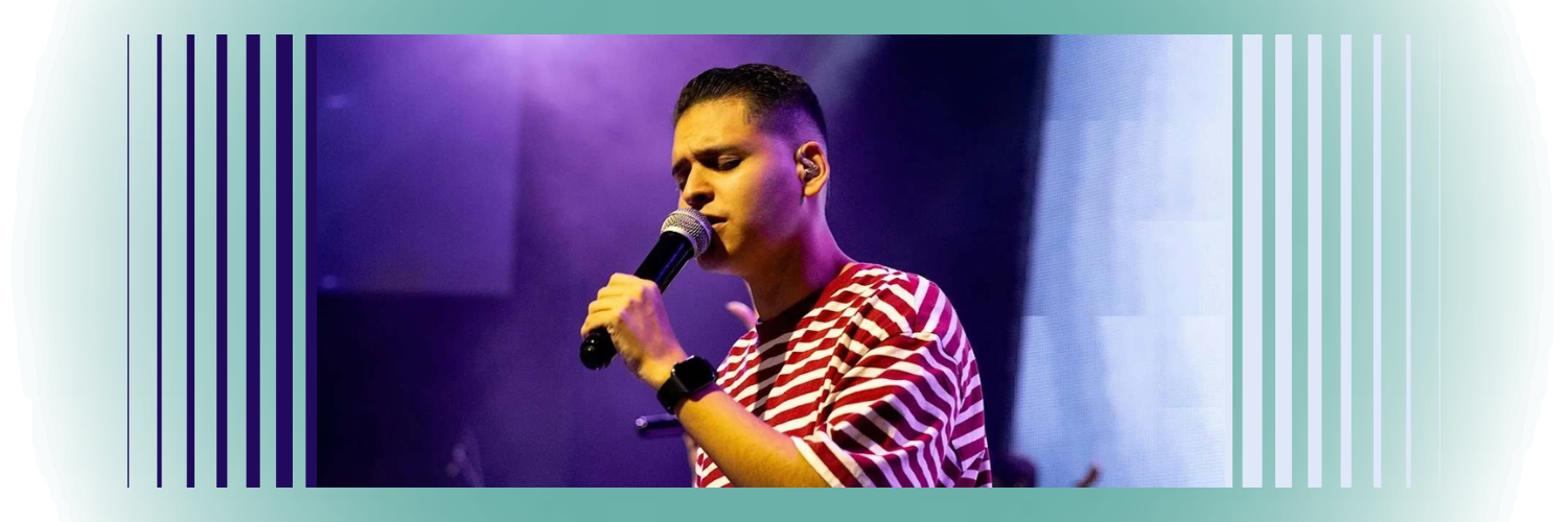 Image shows a young man speaking into a microphone with intense purple lighting behind him.