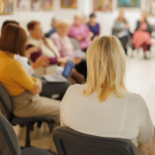 Photograph showing seated adults gathered in a bright room, viewed from behind a blonde person in a white shirt. Group discussion setting includes chairs, colourful clothing, framed artwork, and participants focused toward centre.