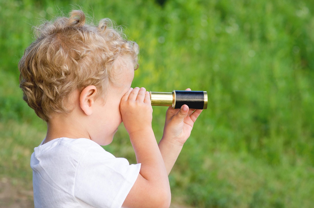 Photograph shows young child using a small brass-and-black telescope outdoors. Curly-haired child in white shirt peers toward greenery, illustrating exploration and observation.