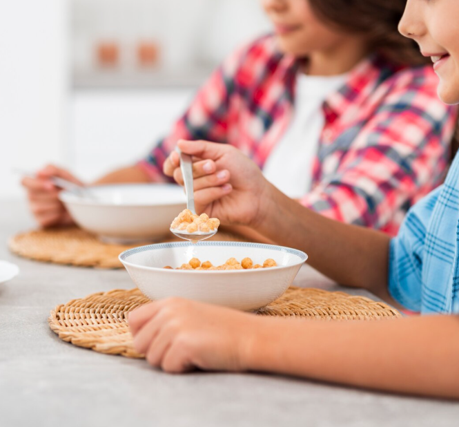 Photograph showing children sharing breakfast at a kitchen table. One child in blue shirt holds a spoonful of cereal above a white bowl, while the other in red plaid shirt eats from another bowl on woven placemats.