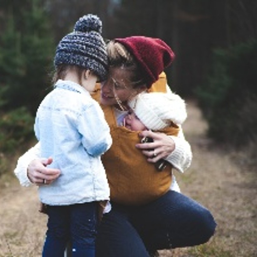 Photograph shows a person kneeling outdoors, embracing two young children among evergreen trees. Children wear winter hats and coats, while the adult wears a red knit hat and mustard sweater, creating a warm family moment in a forest setting.