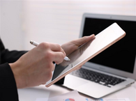 The hands of an unseen person hold a tablet while writing on the screen with an electronic instrument, with a laptop open on a desk in the background.