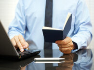 A man sitting at a desk, and visible from waist up to shoulders, types on a laptop keyboard with one hand while holding a passport in the other.