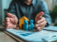 A man shields a model house and figurines with his hands.