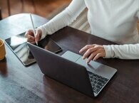 A woman sitting at a desk, visible from her neck down, touches a laptop keypad with her left hand while using a digital pencil on a tablet screen with her right.