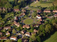 A housing estate viewed from above, encircling a green and with trees and green fields on its fringes.