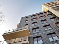 A contemporary block of flats viewed from pavement level with two balconies on the left-hand side and a taller block on the right.