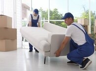 Two men in overalls hold opposite ends of a sofa they're moving, with one standing up and the other crouching down.
