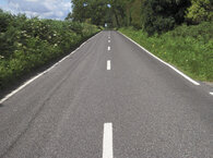 A two-lane tarmac road with white lines down the middle and greenery on either side.